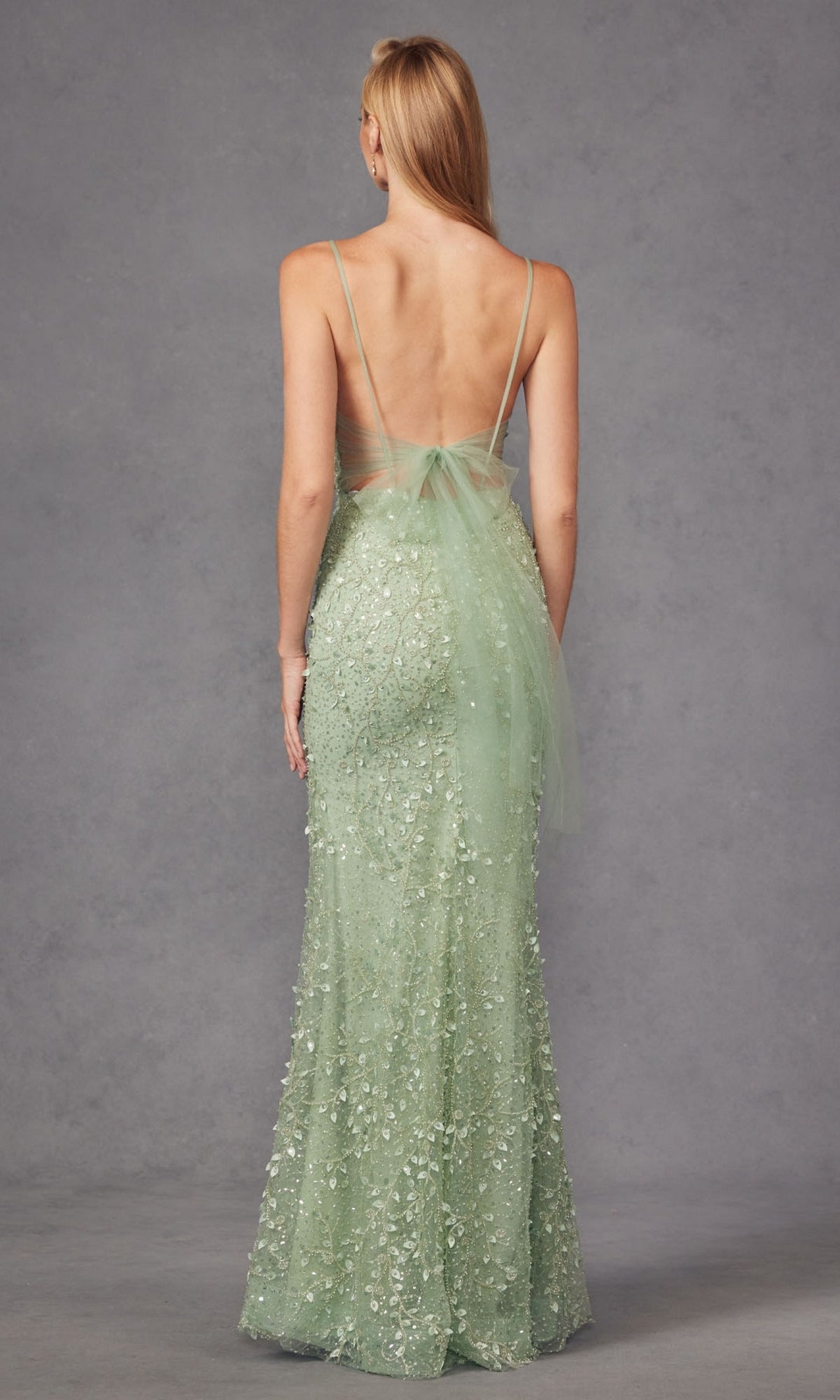 A woman faces away, wearing the Long Prom Dress JT2437A by Juliet in sage green, featuring a plunging v-neckline, backless design, thin straps, and floral embellishments, set against a plain gray background.